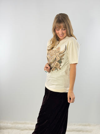 A woman wearing a cream-colored graphic t-shirt with a large floral and horn design on the front, paired with a long black velvet skirt, standing against a light gray studio background.