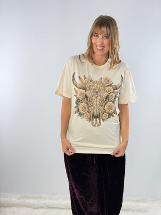 A woman modeling a cream-colored t-shirt with a large graphic of a skull decorated with roses and a crown, paired with dark purple velvet pants, against a light gray studio background.