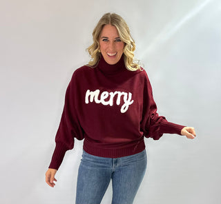 Burgundy loose knit turtleneck sweater with bat sleeves and white monogram "Merry" across the front